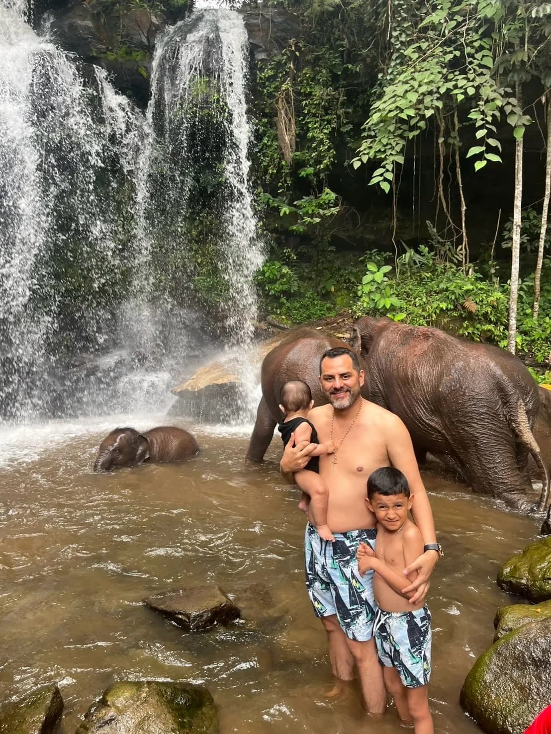 A guest standing waist-deep in the plunge pool with two children while elephants bathe beside them