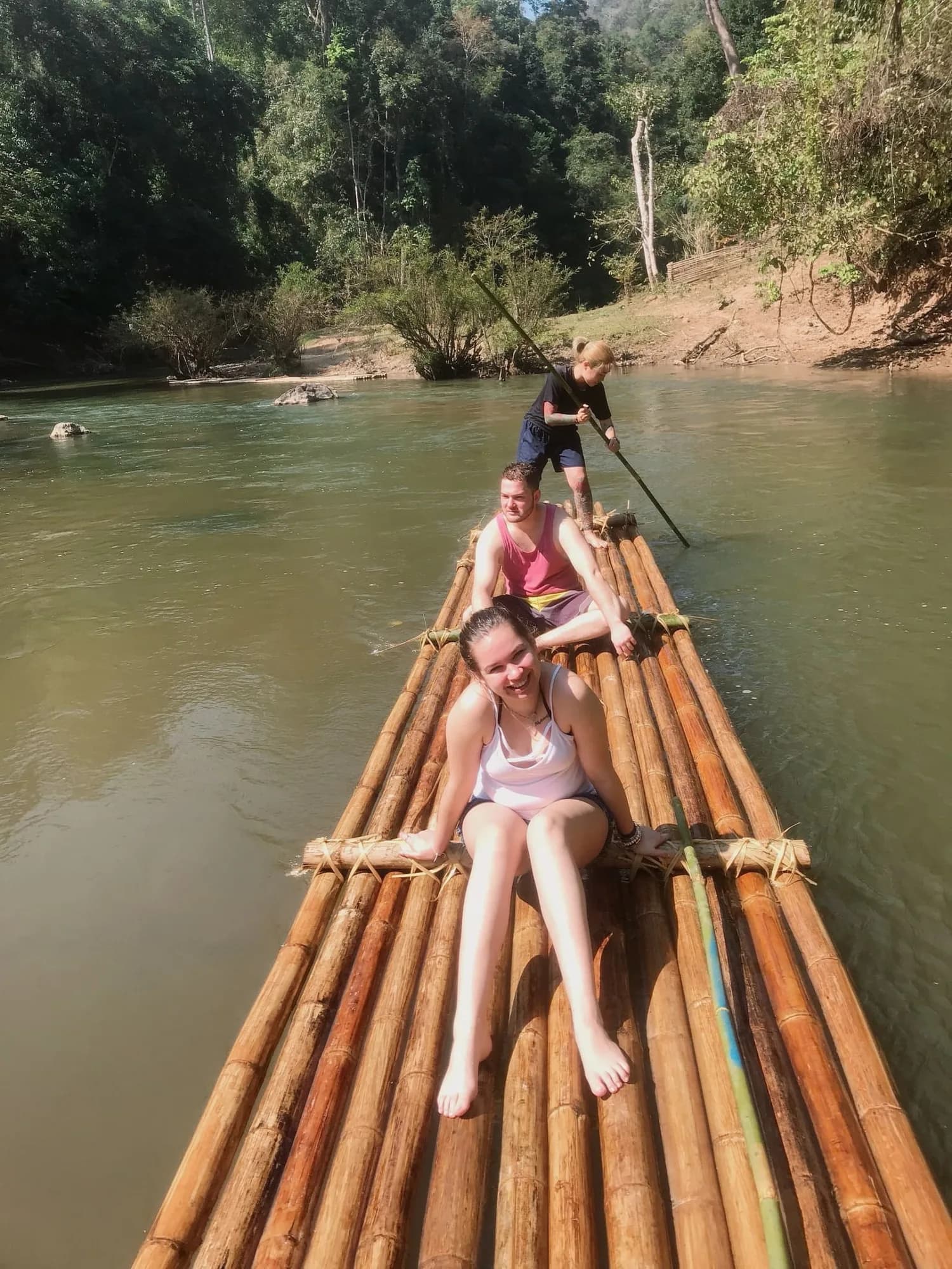 Three guests riding a bamboo raft down a calm green river, one steering with a long pole